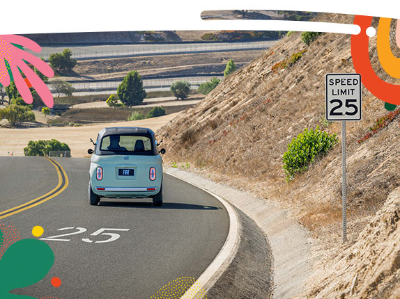 A mint green preproduction model Fiat Topolino traveling down a curved road with a speed limit of 25 miles per hour, shot from a rear angle.