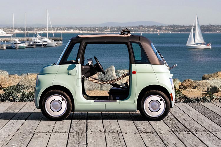 A mint green preproduction model Fiat Topolino Dolce Vita with door ropes and a rolled-up soft top, parked at a marina, shot in profile so you can see through the vehicle and beyond.