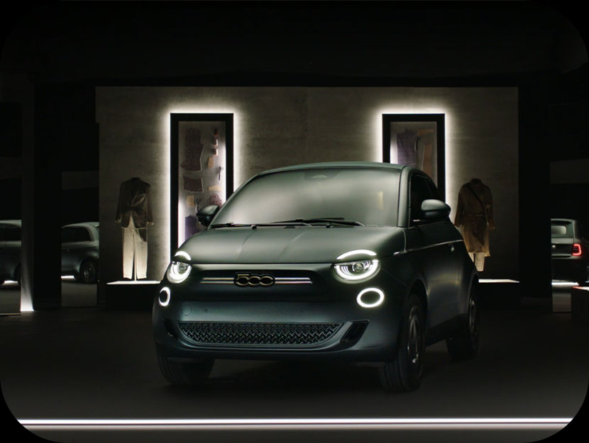 Un Fiat 500e Giorgio Armani Edición Coleccionista negro mate está aparcado en un interior bajo una iluminación espectacular, con faros circulares iluminados y elegantes escaparates de moda al fondo.