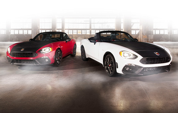 A red 2020 Fiat 124 Spider Abarth and a white 2020 Fiat 124 Spider Abarth with a black hood and its top down, side-by-side in a parking garage.