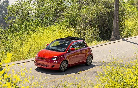 A red European model Fiat 500e being driven on a road surrounded by trees and bushes, with a river and high-rise buildings in the distance.