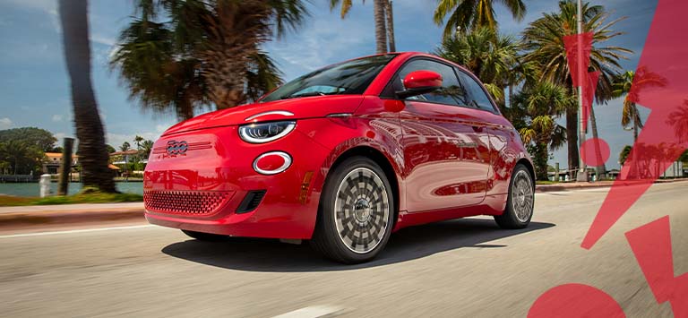 Ángulo delantero del lado del conductor de un Fiat 500e Red 2025 rojo circulando por una carretera con palmeras y un cuerpo de agua al costado, con signos de exclamación ilustrados en rojo, en zigzag, en primer plano, a la derecha.