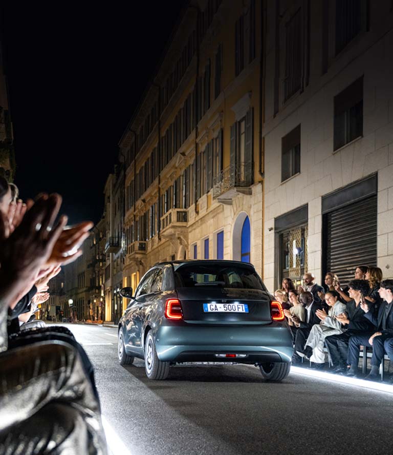 Un ángulo trasero de un FIAT 500e Edición de Colección Giorgio Armani 2025 verde oscuro circulando por una calle dispuesta como una pista iluminada, pasando junto a un grupo de espectadores.