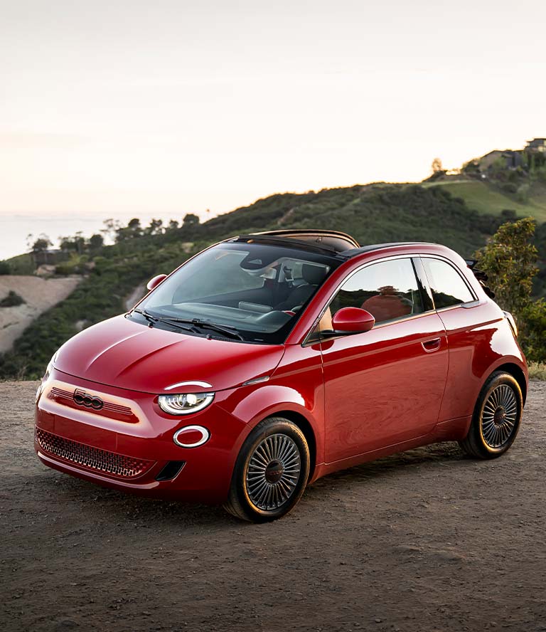 Perfil frontal en ángulo, del lado del conductor, de un Fiat 500e rojo, modelo europeo, de 2024, con el techo corredizo abierto, estacionado en un claro con montañas y el mar de fondo.